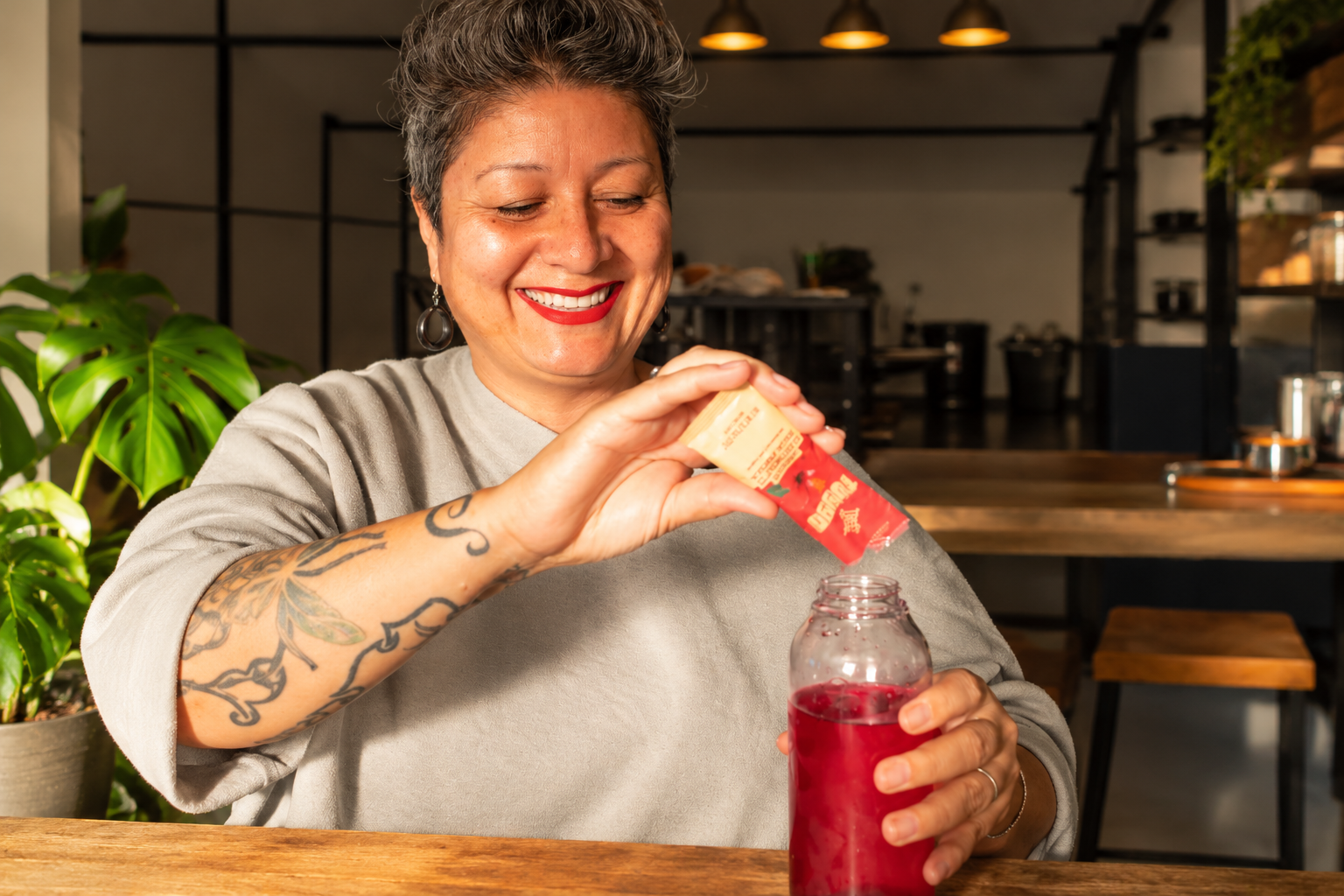 A smiling woman pours a red Tuyyo Foods Agua Fresca drink mix into a clear bottle indoors.