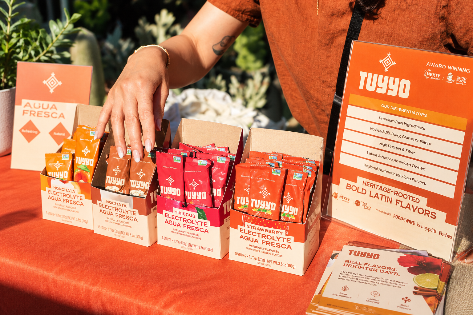Tuyyo Foods Agua Fresca sachets and branded display cartons are arranged on an orange table at an outdoor event.