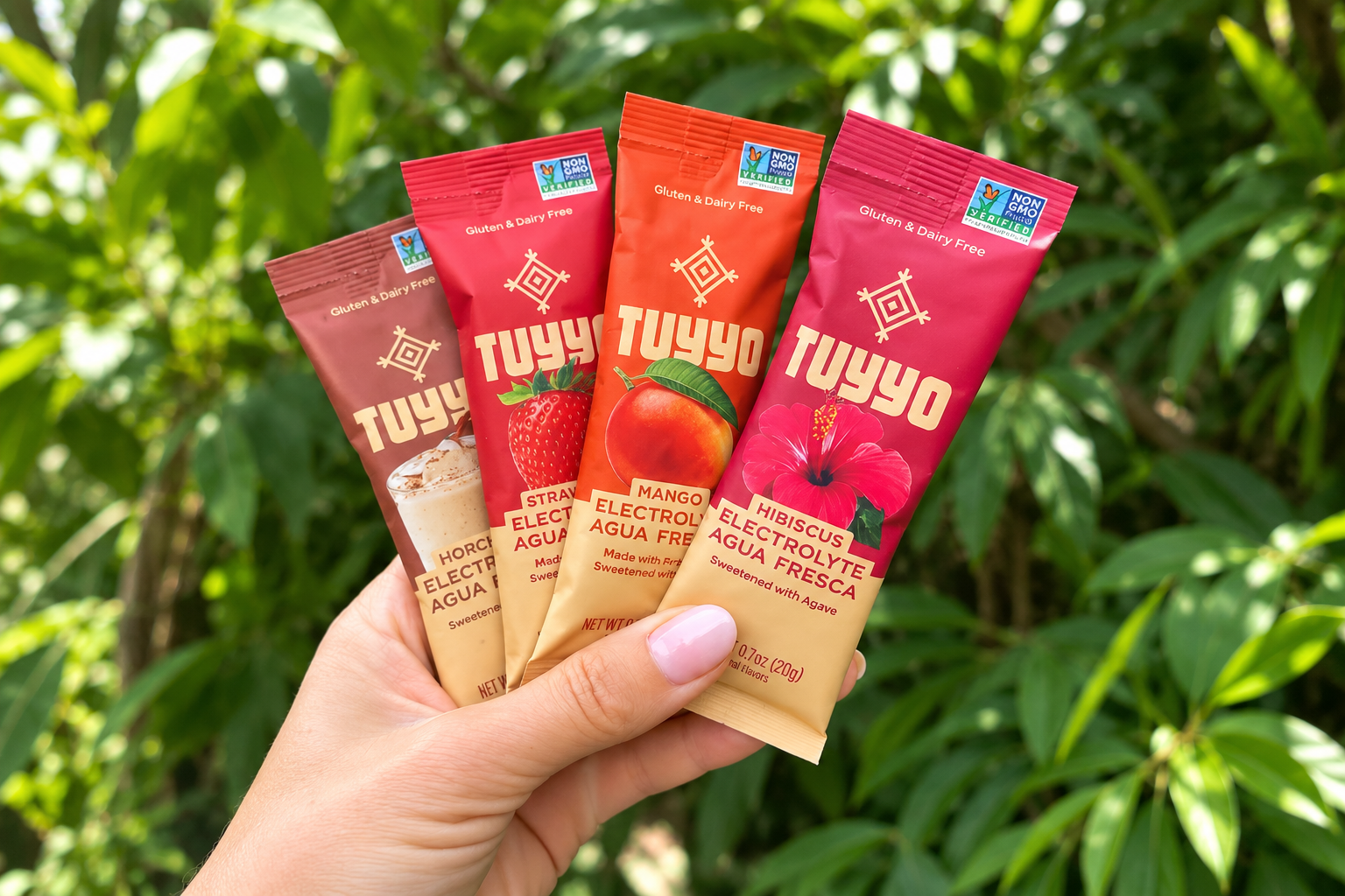 A hand holds four colorful Tuyyo Foods electrolyte Agua Fresca sachets against green foliage.