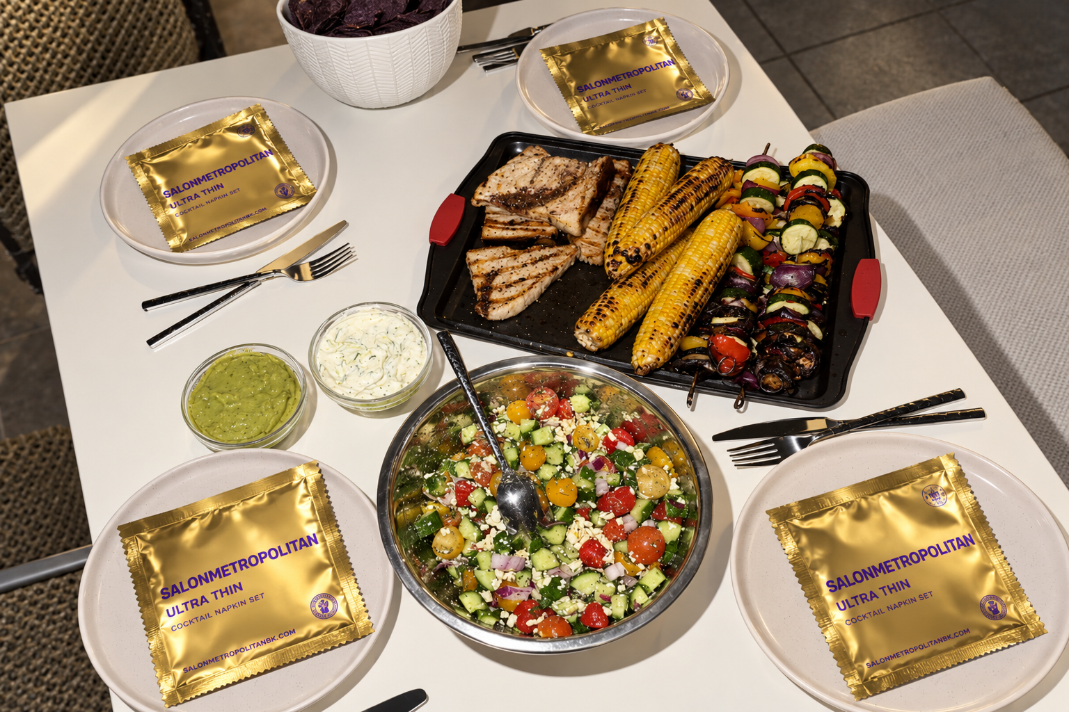 Gold Salon Metropolitan cocktail napkin sachets sit on plates around an outdoor dining table with grilled food and salads.