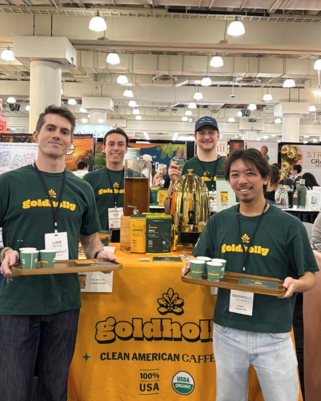 Four people in green Goldholly shirts at a trade-show booth, with tea pouches and serving trays.