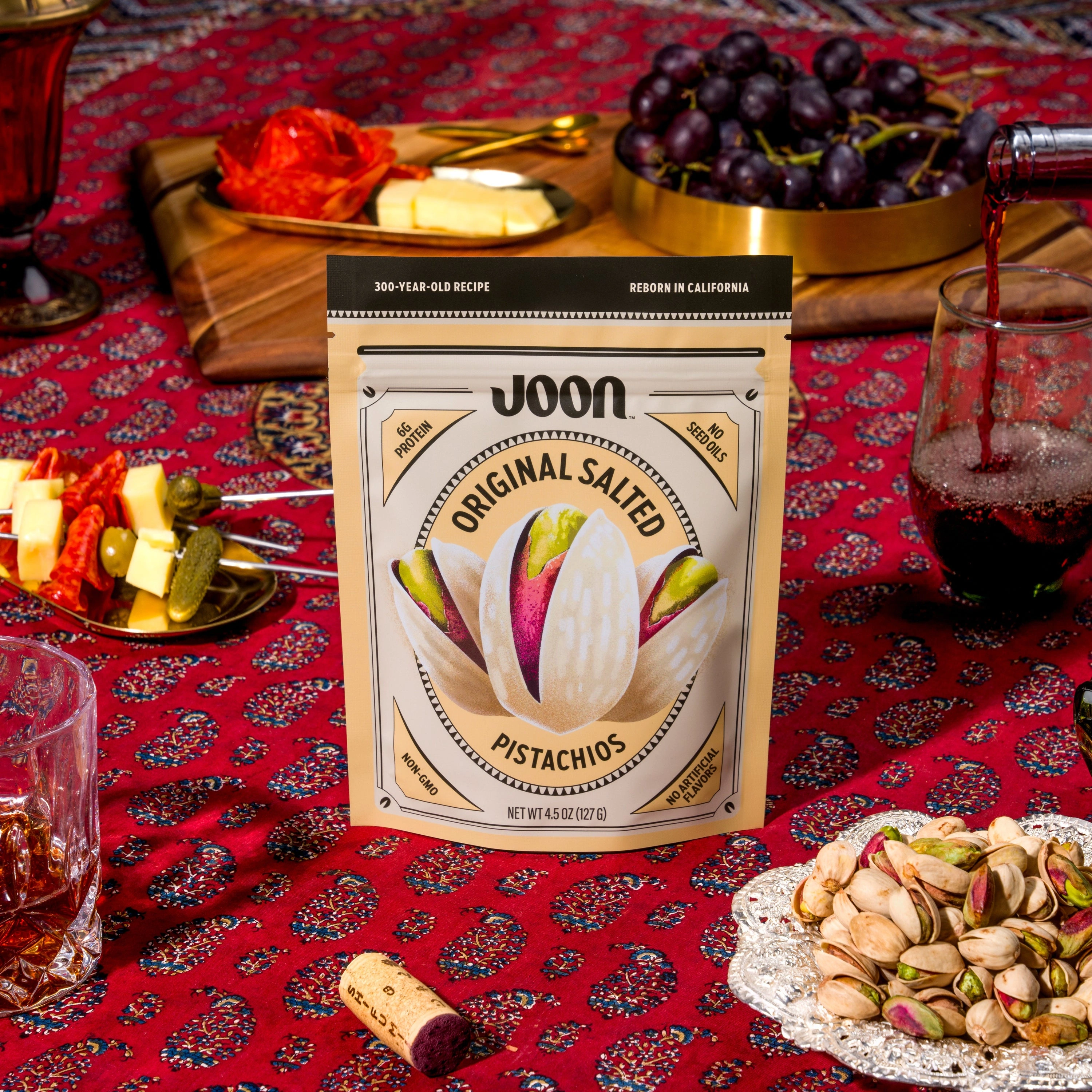 Joon Original Salted Pistachios pouch on a red patterned table with grapes, cheese, pistachios, and a poured drink.