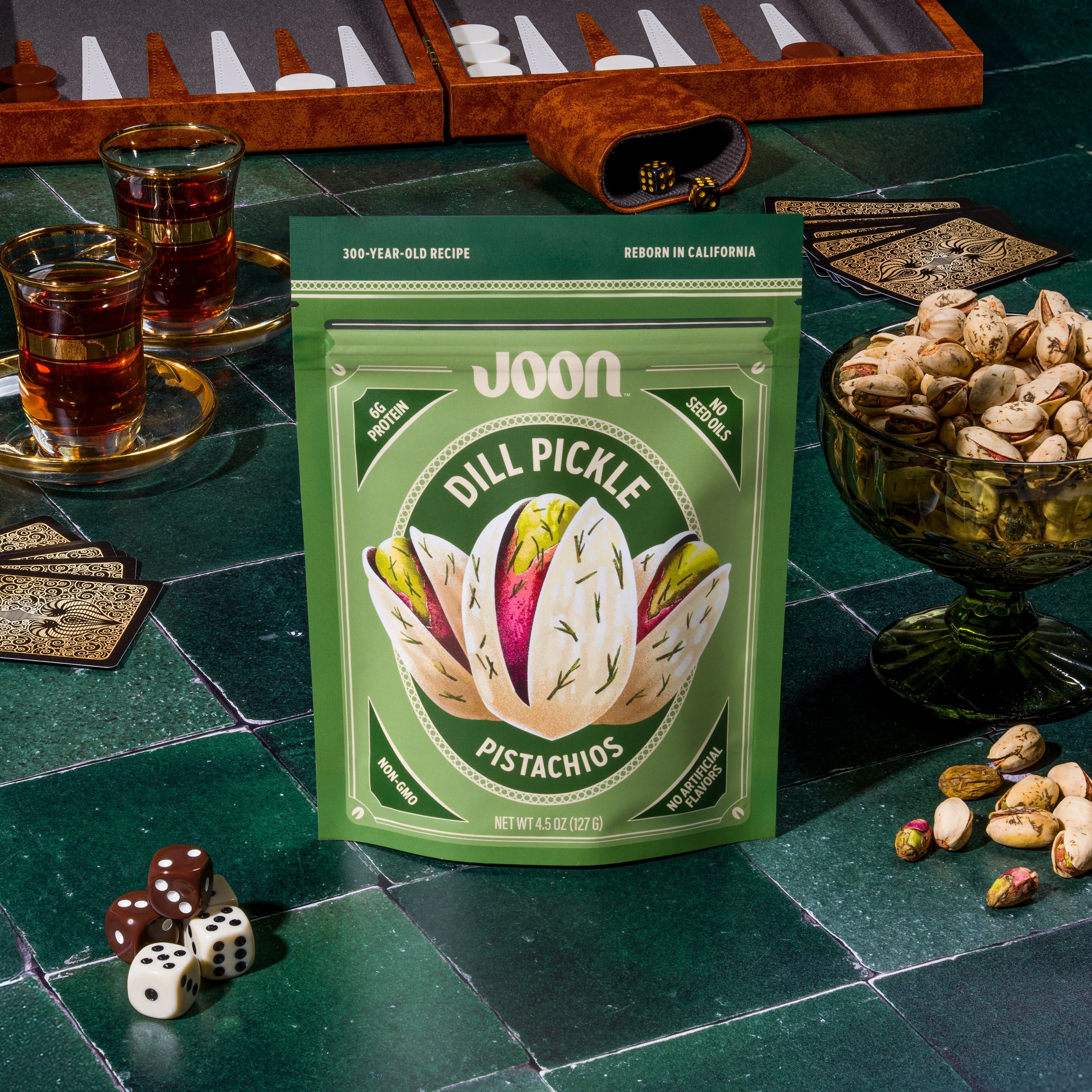 Joon Dill Pickle Pistachios pouch on a green game table with backgammon, cards, dice, tea, and pistachios.