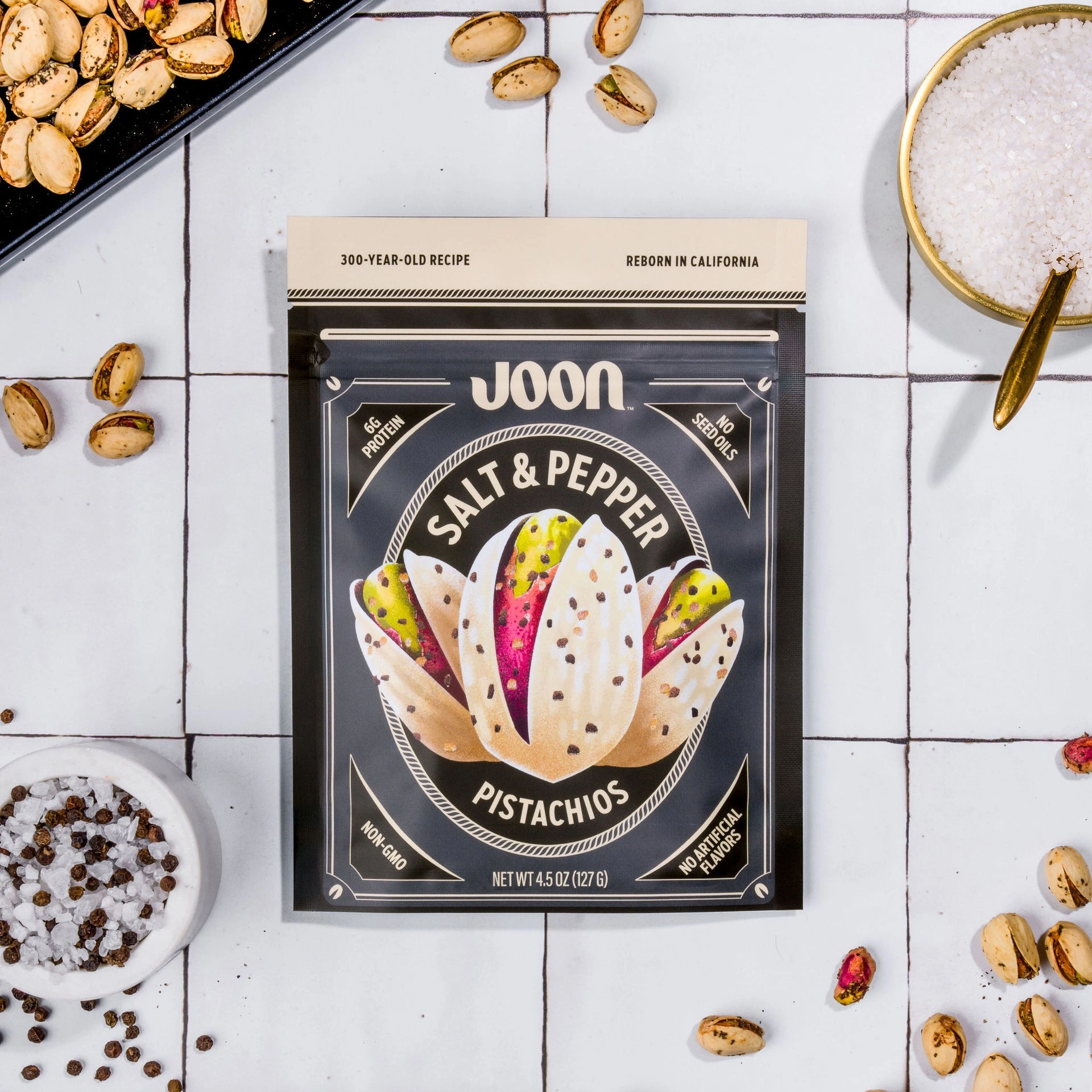 Joon Salt & Pepper Pistachios pouch on a white tiled surface with seasoned pistachios, salt, and pepper.