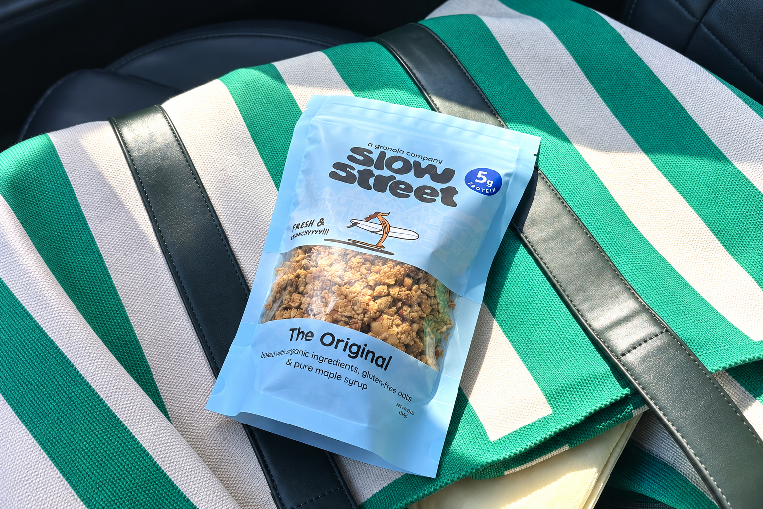 A light blue Slow Street Granola Original pouch rests on a green and white striped tote bag.