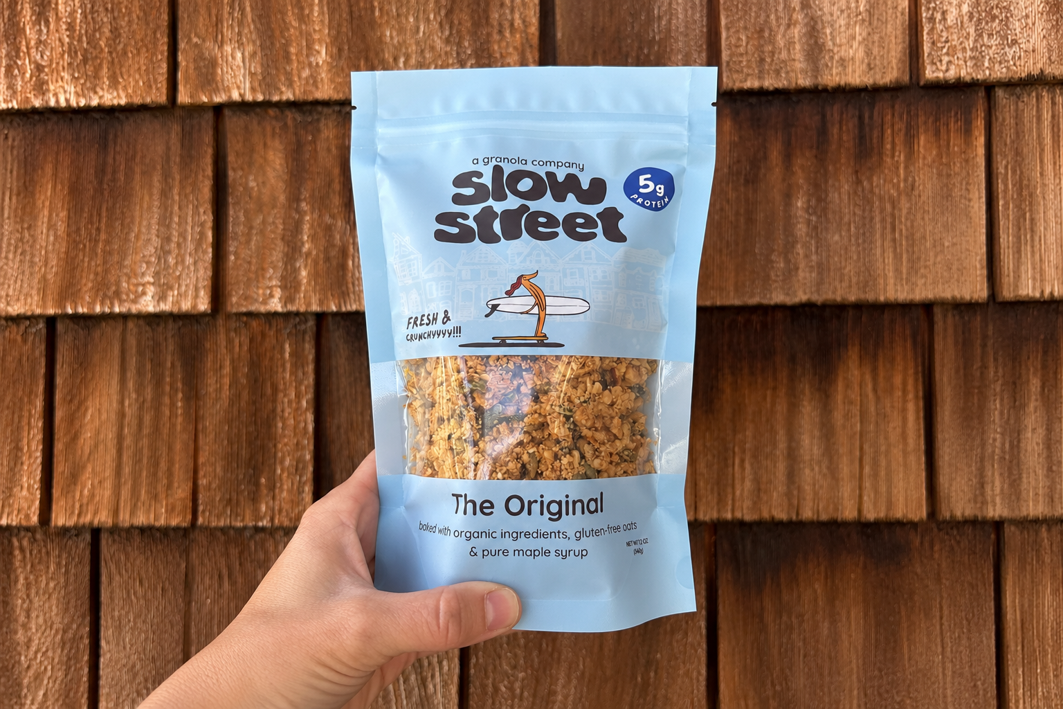 A hand holds a light blue Slow Street Granola Original pouch against a wooden shingle wall.