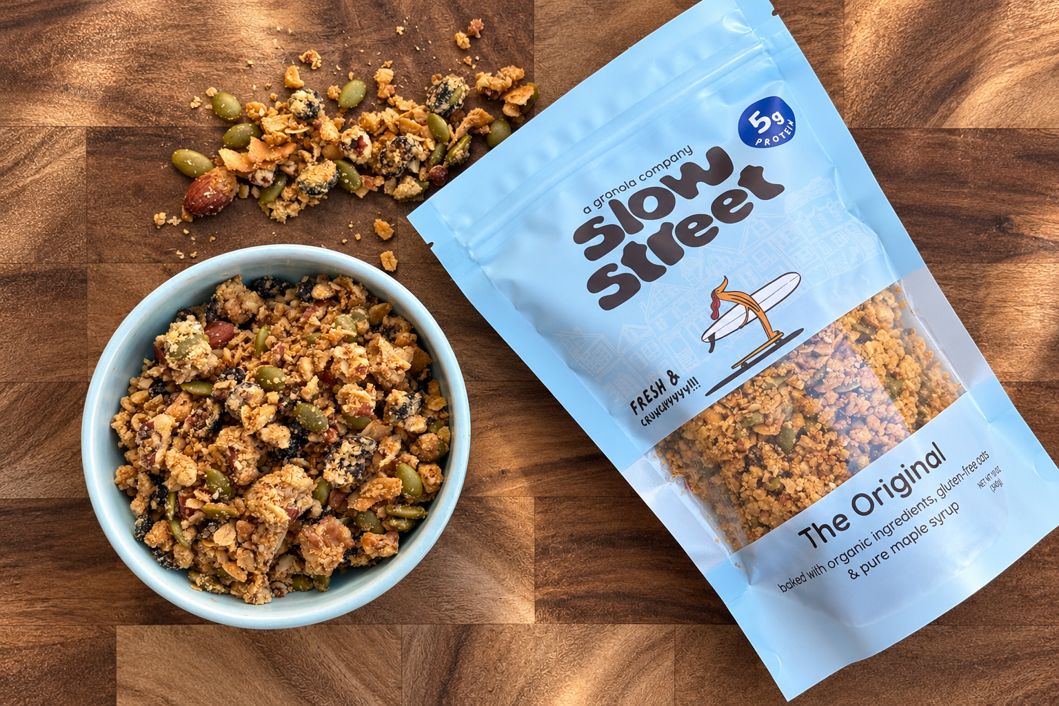 A light blue Slow Street Granola Original pouch sits beside a bowl of granola on a wooden surface.