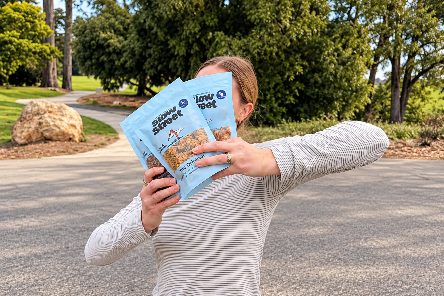 A woman holds three light blue Slow Street Granola pouches in front of her face outdoors.
