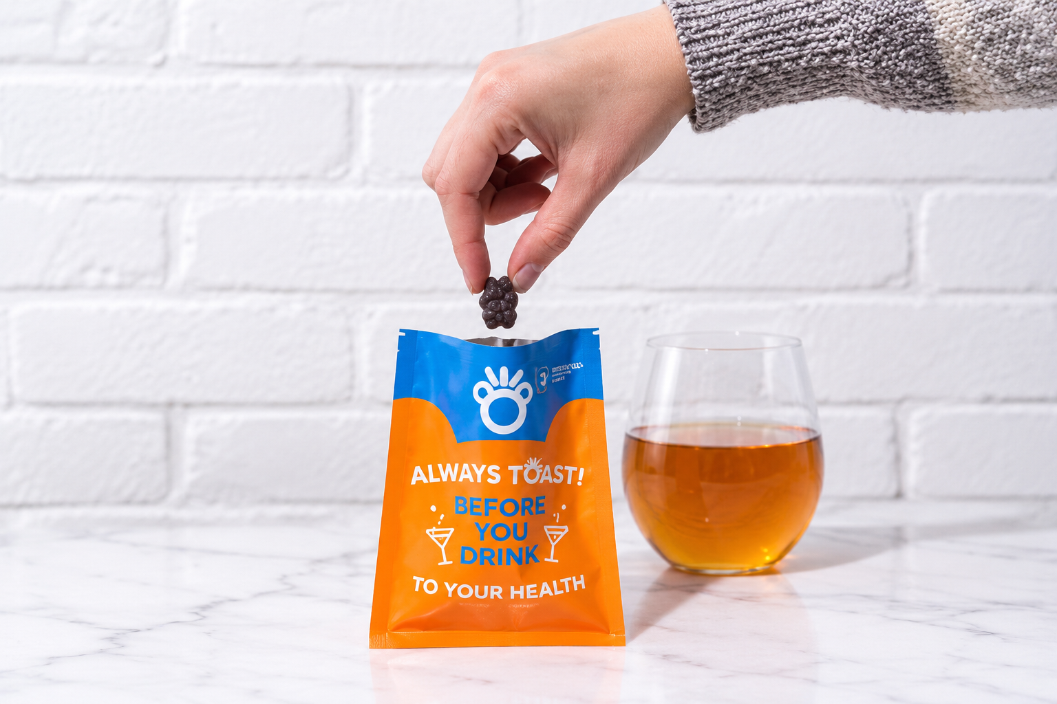 A hand holds a dark gummy over an open Toast! supplement pouch beside a glass of amber-colored drink.