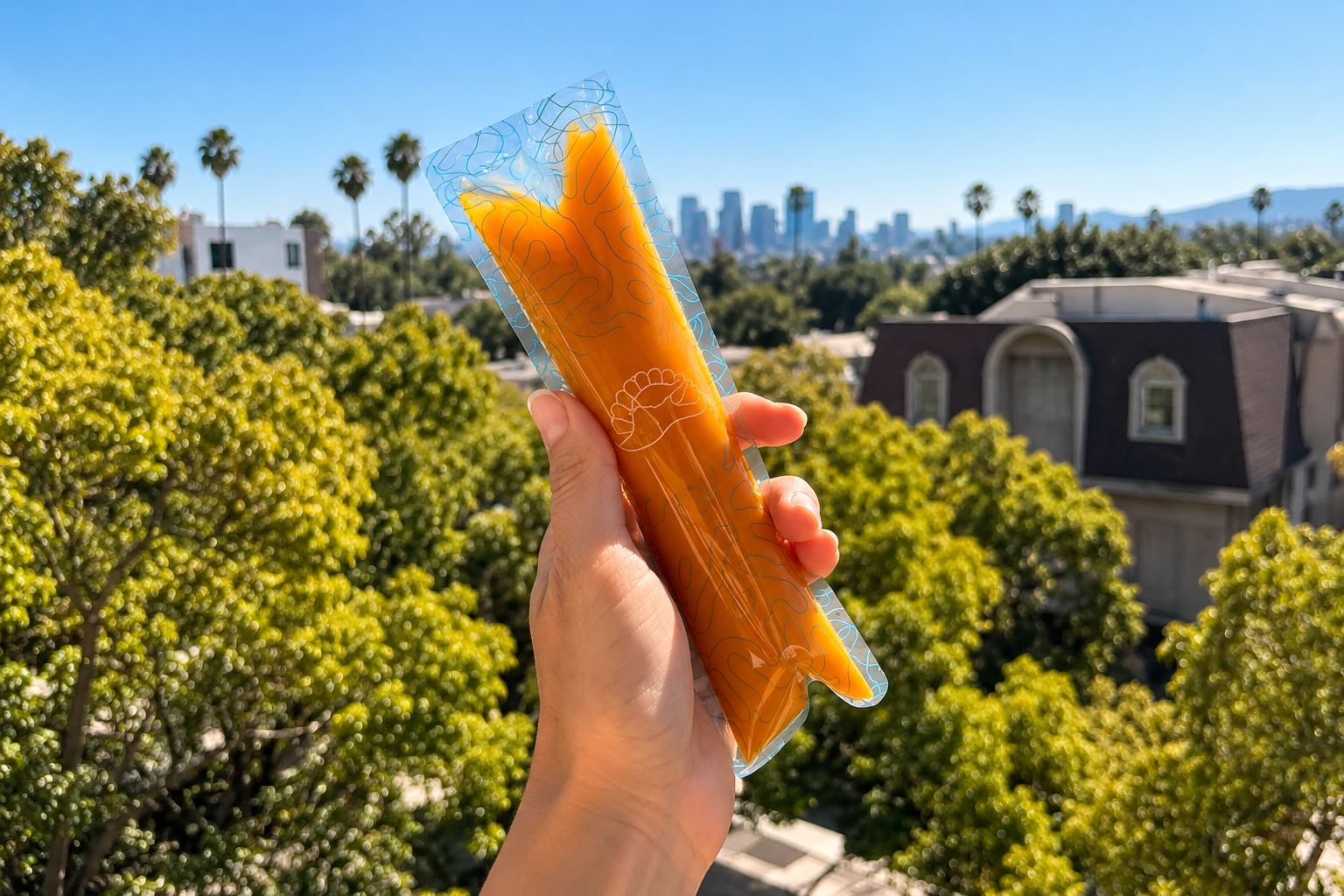 A hand holds an orange Sea Gems popsicle above trees with the Los Angeles skyline in the distance.