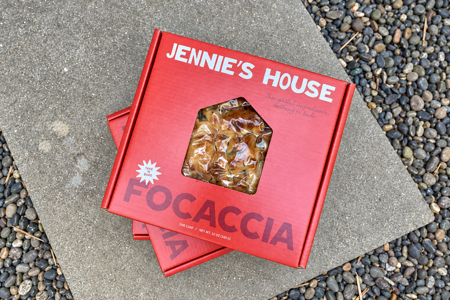 Two red Jennie's House focaccia cartons rest on a stone surface beside smooth pebbles.