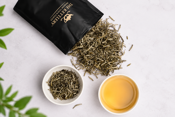 A black The Kettlery tea pouch, loose leaves, a bowl of tea, and a cup are arranged on a white surface.