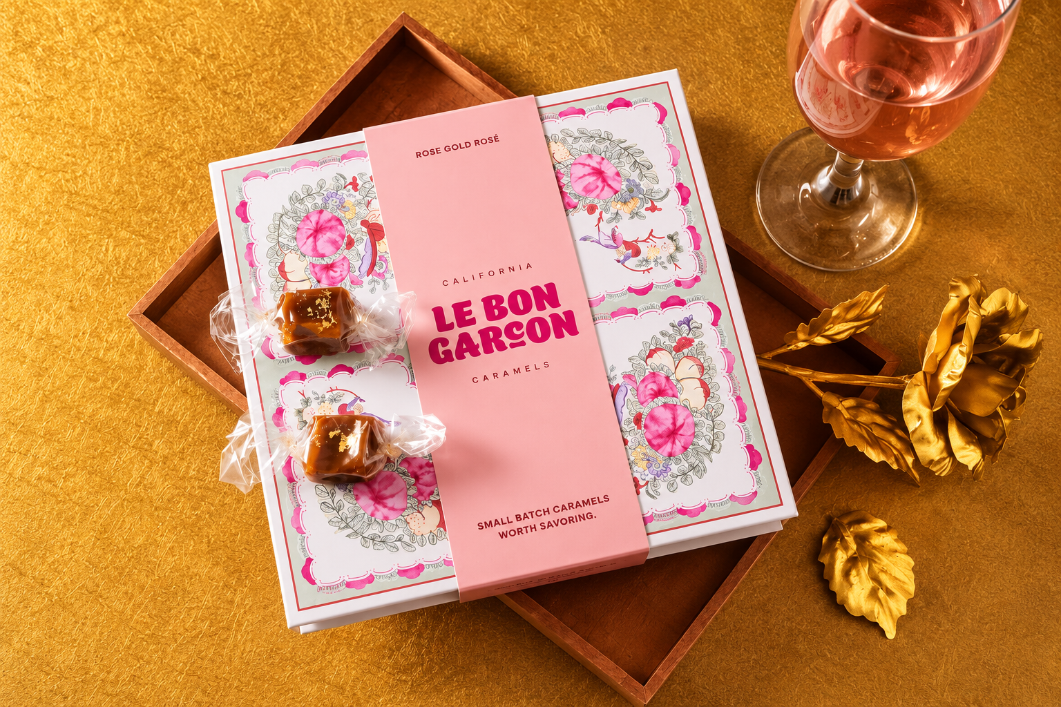 A pink Le Bon Garçon rigid box with floral artwork is displayed with wrapped caramels and a glass of rosé.